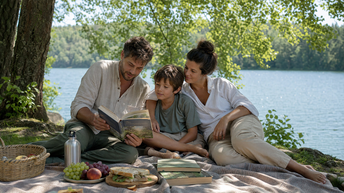 Eine Pflegefamilie beim entspannten Picknick an einem See im Sommer, ein Elternteil liest dem Kind aus einem Buch vor.