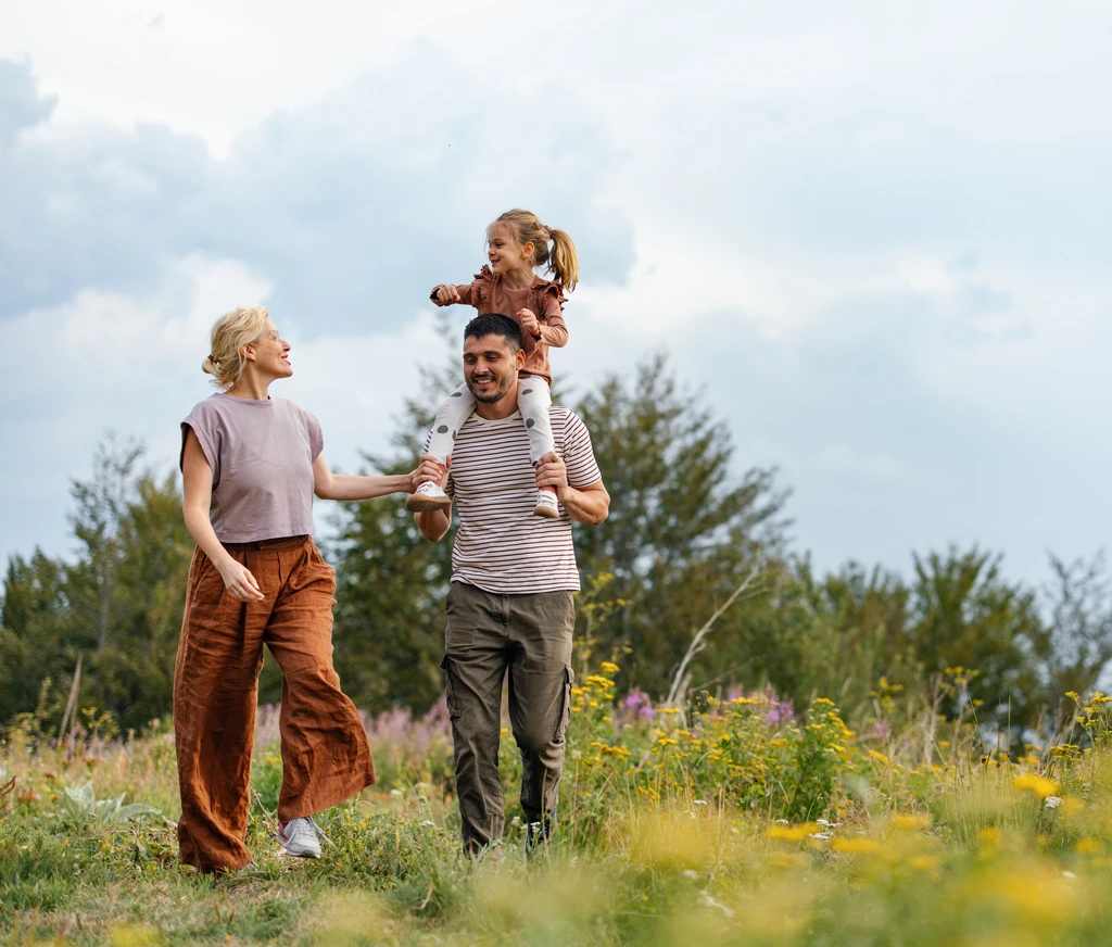 Pflegeeltern sind mit ihrem Pflegekind in der Natur unterwegs, der Vater trägt das Mädchen auf den Schultern.